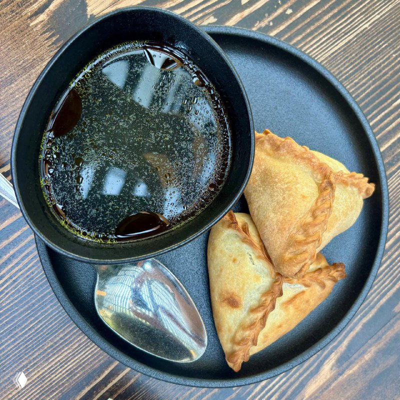 Вид сверху: тёмная миска с бульоном, рядом две треугольные лепёшки/пирожка и ложка на тёмной тарелке — сервировка в ресторане.