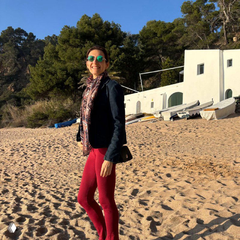 Женщина на песчаном пляже в солнечных очках и шарфе, улыбается на фоне белых домов и прибрежных сосен