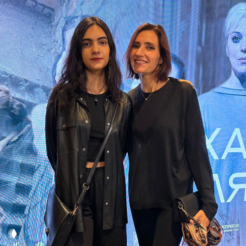 Две женщины в тёмной одежде позируют на фоне подсвеченного экрана; фото сделано на мероприятии, спокойная дружелюбная атмосфера.