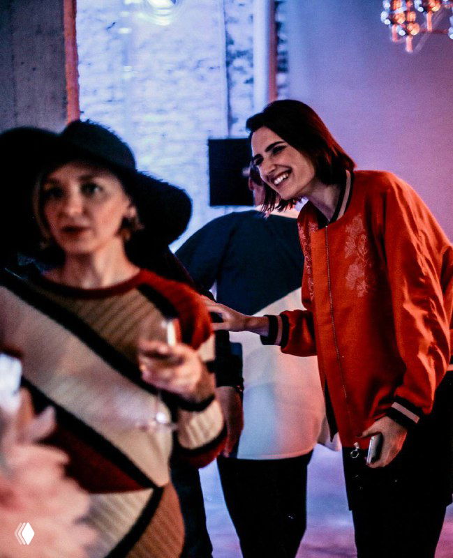 Улыбающаяся женщина в красной куртке на светском мероприятии; на переднем плане размытая дама с бокалом