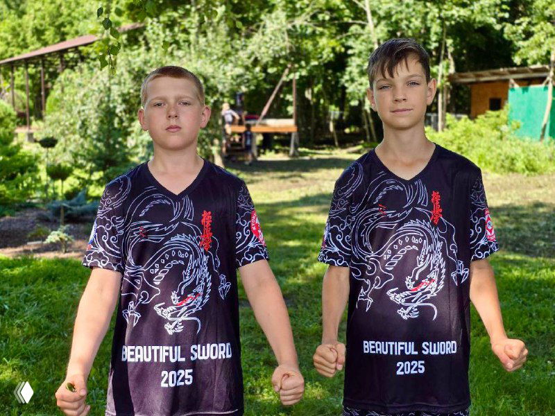 Двое мальчиков в одинаковых футболках лагеря карате стоят на зелёной поляне лагеря среди деревьев, на заднем плане палатки и спортивная площадка.