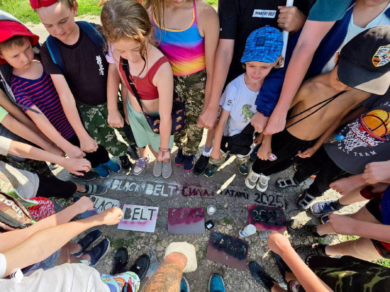 Группа детей и подростков в спортивной форме собраны в круг и смотрят на мелом расписанную землю, общий момент лагерного единения и командного духа.