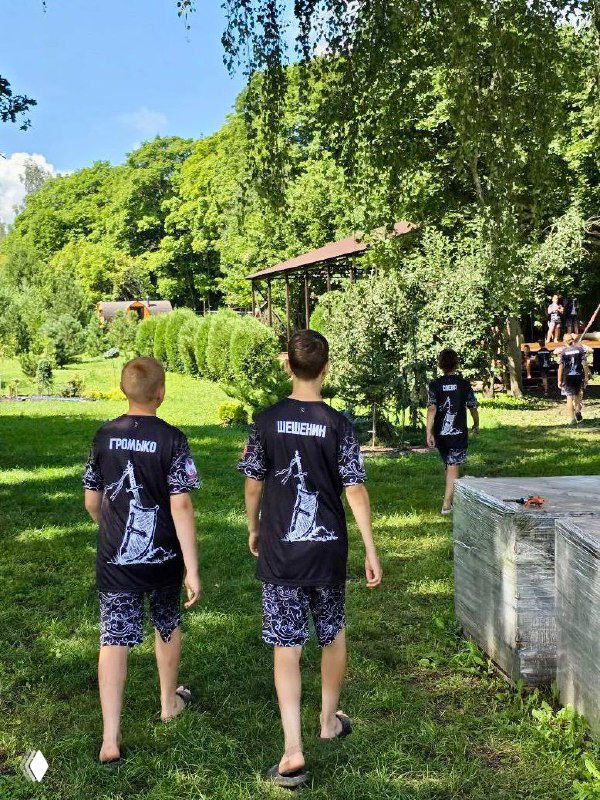 Двое мальчиков в футболках лагеря идут по тропинке к спортивной поляне и беседке, видны элементы лагерной инфраструктуры и другие участники сборов.