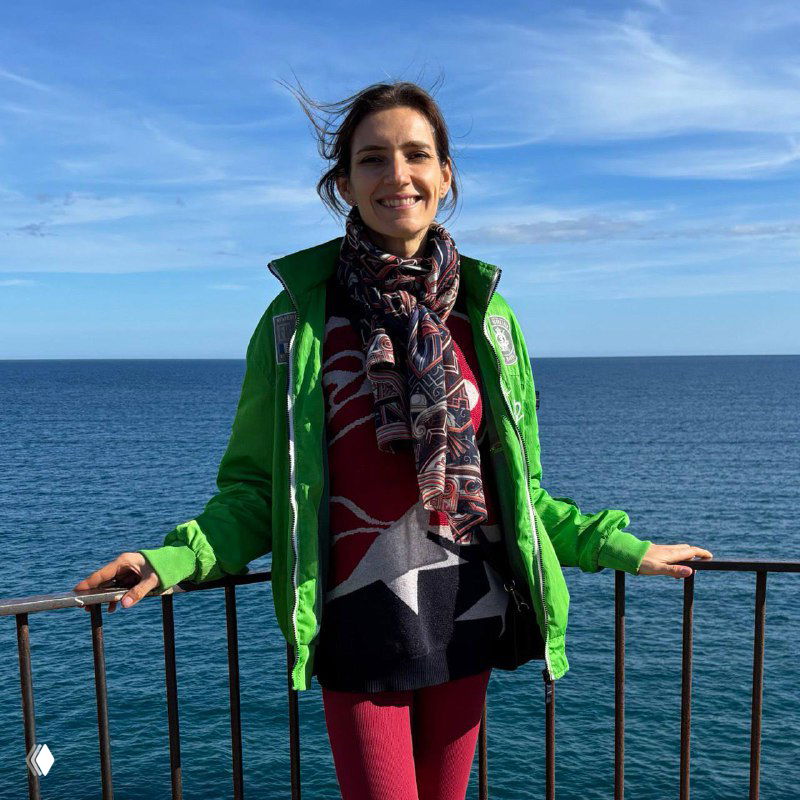 Женщина в зелёной куртке улыбается на фоне спокойного моря, стоит у ограждения на набережной — портрет авторки в естественной обстановке.