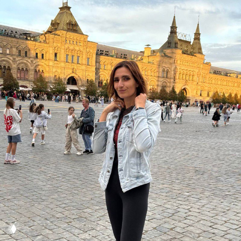 Женщина стоит на широкой городской площади на фоне исторического фасада и прохожих — иллюстрация о формировании уверенности и практики.