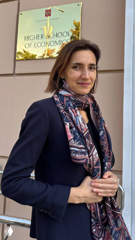 Женщина на фестивале школьников у таблички «Высшая школа экономики» в пальто и шарфе у входа университета, деловой городской фон и логотип мероприятия на заднем плане.