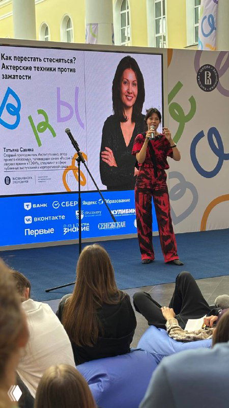 Лектор выступает на сцене перед аудиторией школьников и студентов: большая проекция с фотографией спикера, сидящие слушатели и атмосфера образовательного события в ВШЭ.