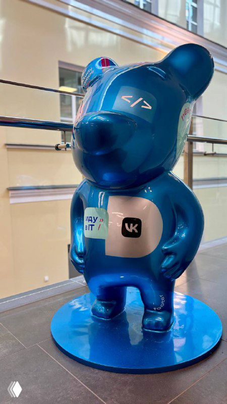 Синяя декоративная статуэтка в виде медвежонка с логотипами и стикерами IT/платформ, установленная в зоне фестиваля и служащая элементом оформления и фотозоны мероприятия.