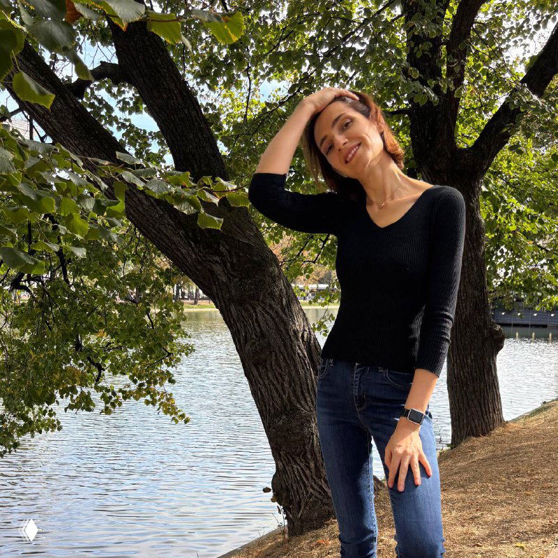 Виктория Шешенина стоит у реки под деревом в повседневной одежде, улыбается в камеру — портретный снимок на открытом воздухе.