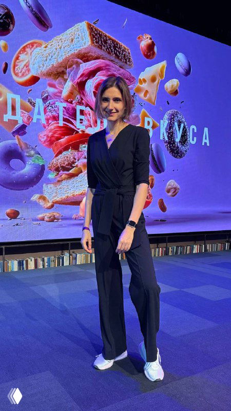 Виктория Шешенина стоит на сцене в деловом костюме, улыбается на фоне большого яркого слайда с десертами.