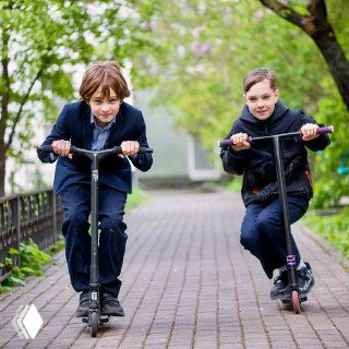 Два мальчика на самокатах катаются по дорожке у школьного здания — активные перемены и детская мобильность на территории образовательного учреждения.