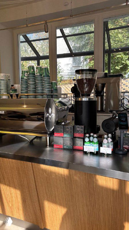 Кофемолка, оборудование и одноразовые стаканчики на прилавке: рабочая зона баристы с деталями приготовления напитков и упаковками воды.