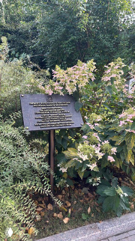 Табличка или мемориальная доска у зелёной зоны парка возле кафе: информационный знак на фоне кустов и декоративных растений вдоль тропинки.