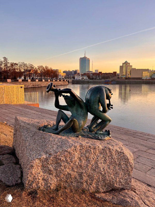 Скульптура у набережной Челябинска на фоне воды и городской застройки при тёплом вечернем небе и спокойной воде.