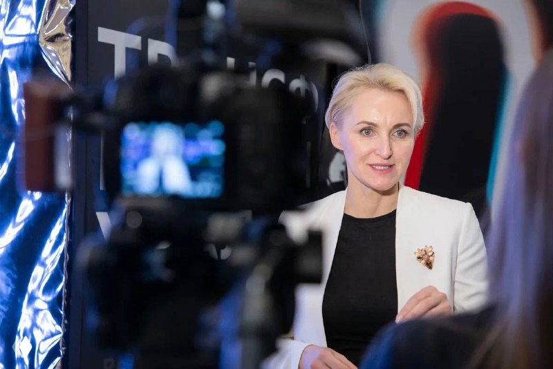 Женщина даёт интервью на фоне бренд‑волла форума: камера на переднем плане, спикер ярко освещена и говорит в кадр.