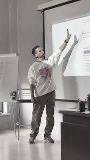 Лектор у экрана слайдов в светлом зале: мужчина в повседневной одежде указывает на проекцию с презентацией — иллюстрация мастер‑класса по соцсетям и автоматизации.