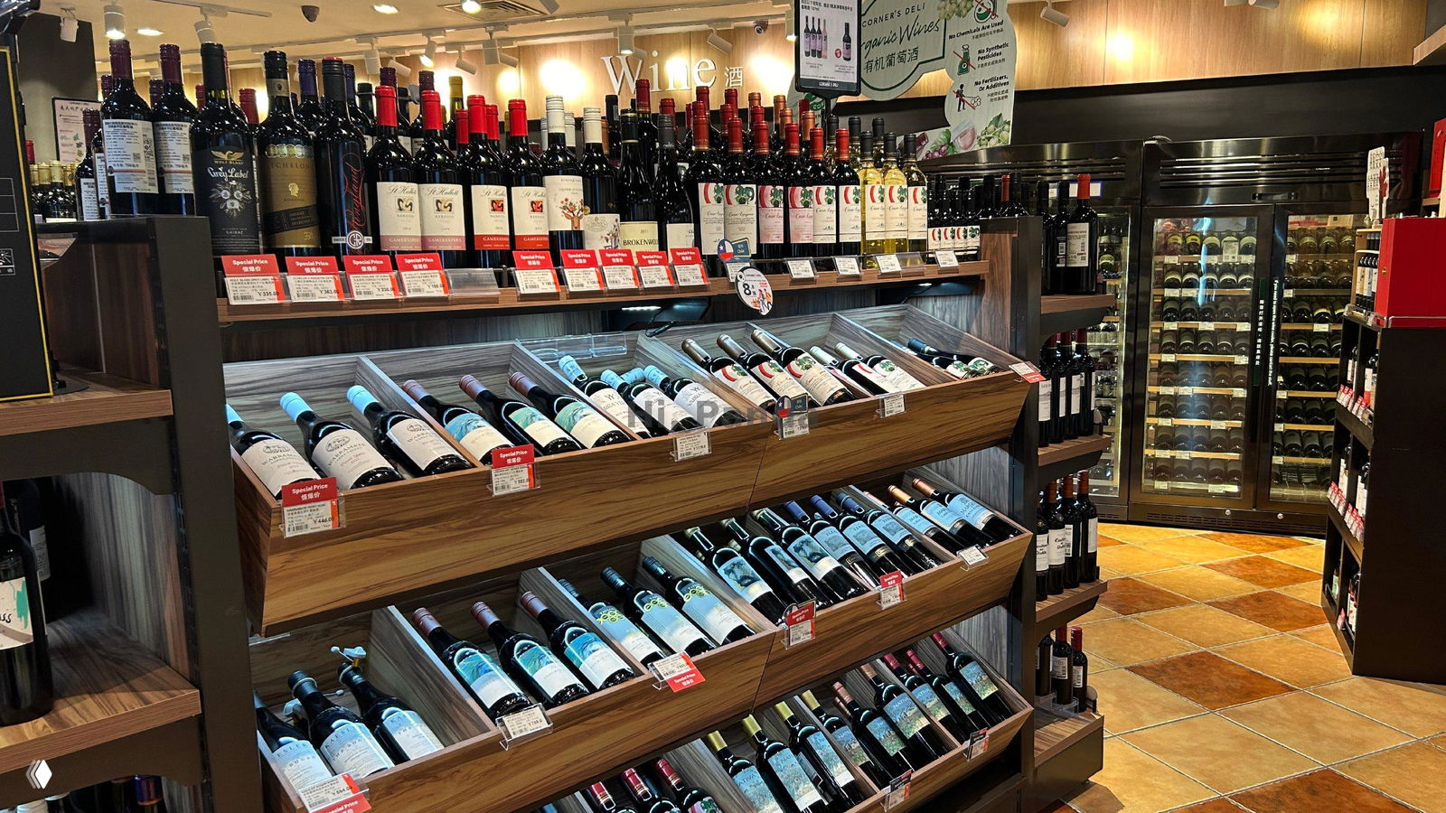 Деревянные наклонные лотки и полки с бутылками вина, ценники на передней панели и охлаждающий шкаф на заднем плане в магазине ТЦ «Лето».