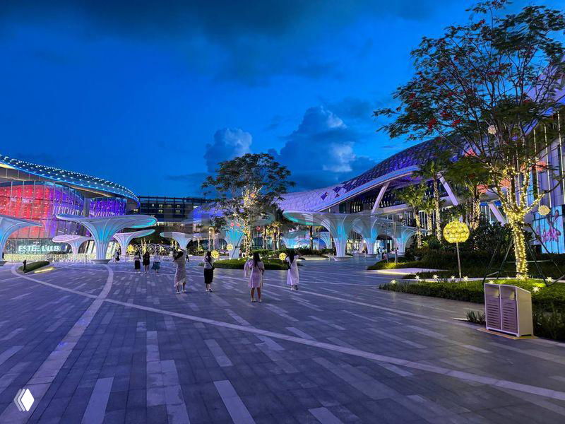 Ночная фотография открытой площади у торговой зоны Хайнаня: освещённые павильоны, пешеходные дорожки и прохожие.