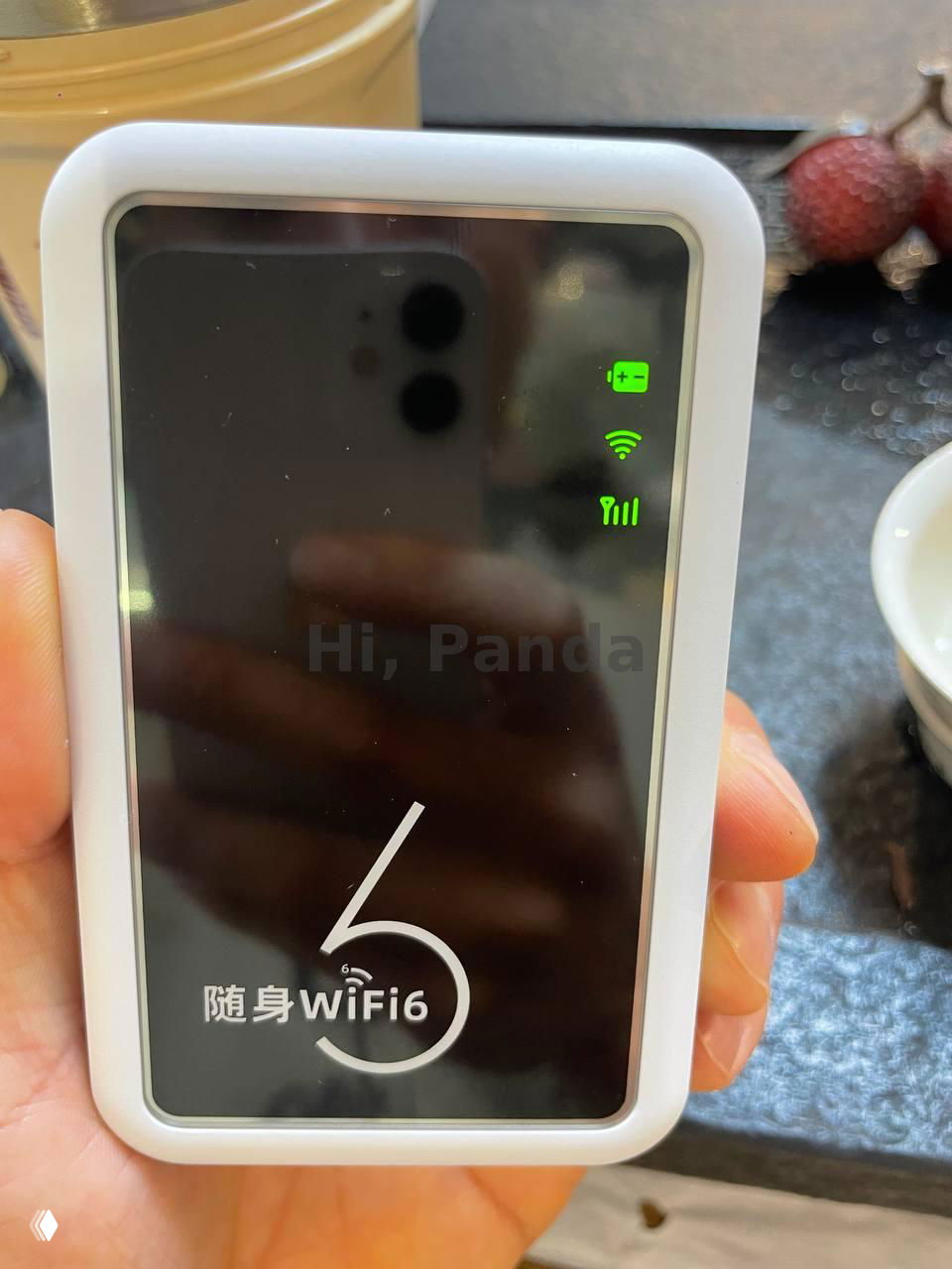 Портативный белый Wi‑Fi роутер с надписью «WiFi6», зелёными индикаторами сигнала и заряда, который держат в руке на кухонном фоне.