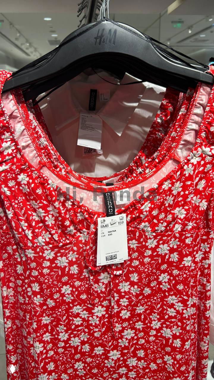 Красное платье или топ в цветочный принт на вешалке H&M; видно пластиковую вешалку с логотипом H&M и ценник в RMB, крупный вид товара на стойке магазина.