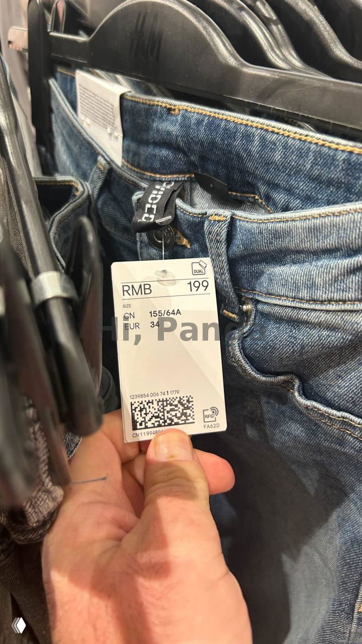 Рука держит бирку джинсов с отметкой RMB 199 внутри магазина; видно швы джинс, ценник и часть торговой стойки в магазине ТЦ Mangrove Tree Sanya.