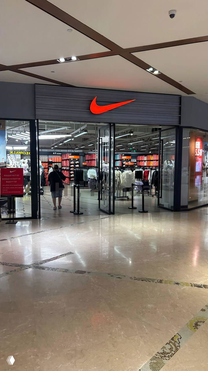 Вход в магазин Nike в торговом центре Mangrove Tree Sanya: фасад с красным логотипом, стеклянные двери и прохожие на полированном полу торгового центра.
