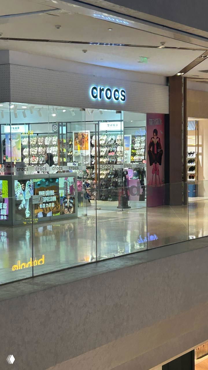 Магазин Crocs в торговом центре: внутренние стеллажи, яркие полки с обувью и вид через стеклянный балкон на уровень ниже.