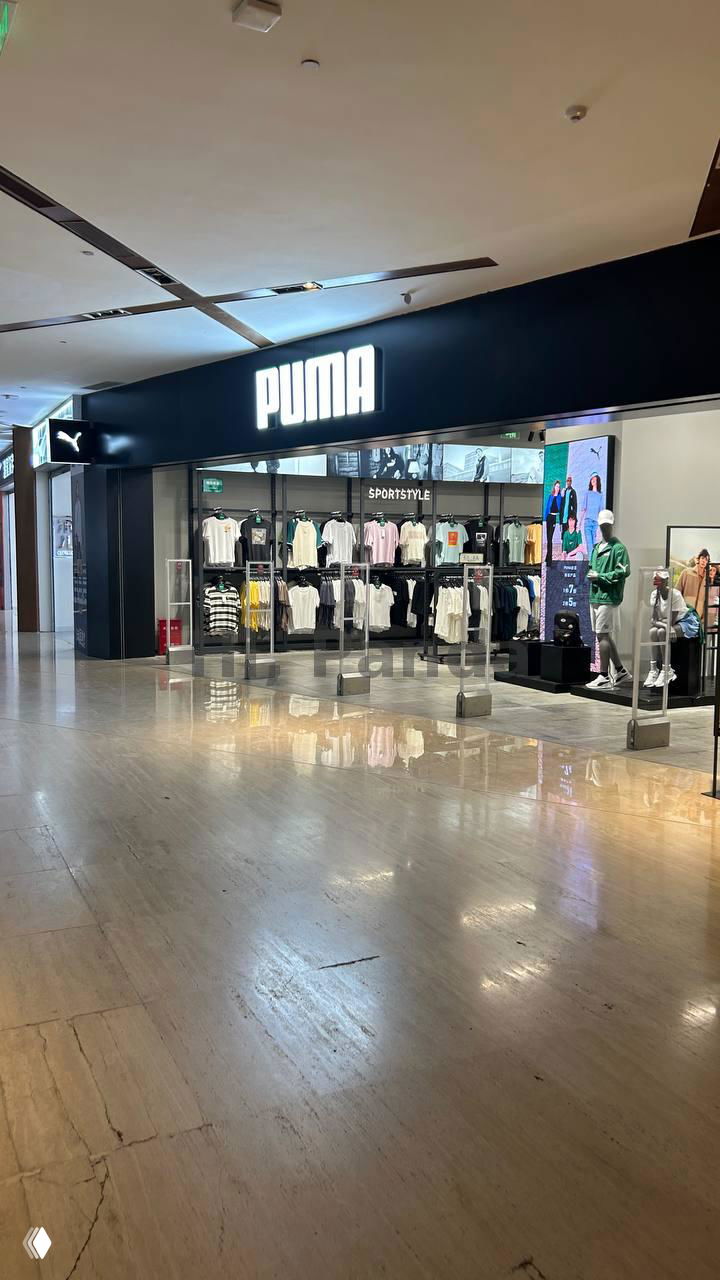 Магазин PUMA с черной вывеской и внутренней выкладкой одежды: просторный вход и отражение света на полированном полу торгового центра.