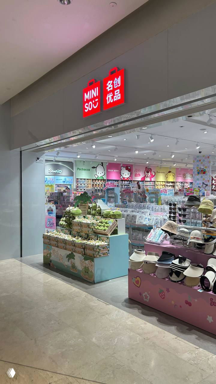 Мини-сеть Miniso с ярким интерьером: полки с аксессуарами, мягкими игрушками и головными уборами в пастельных тонах внутри торгового центра.