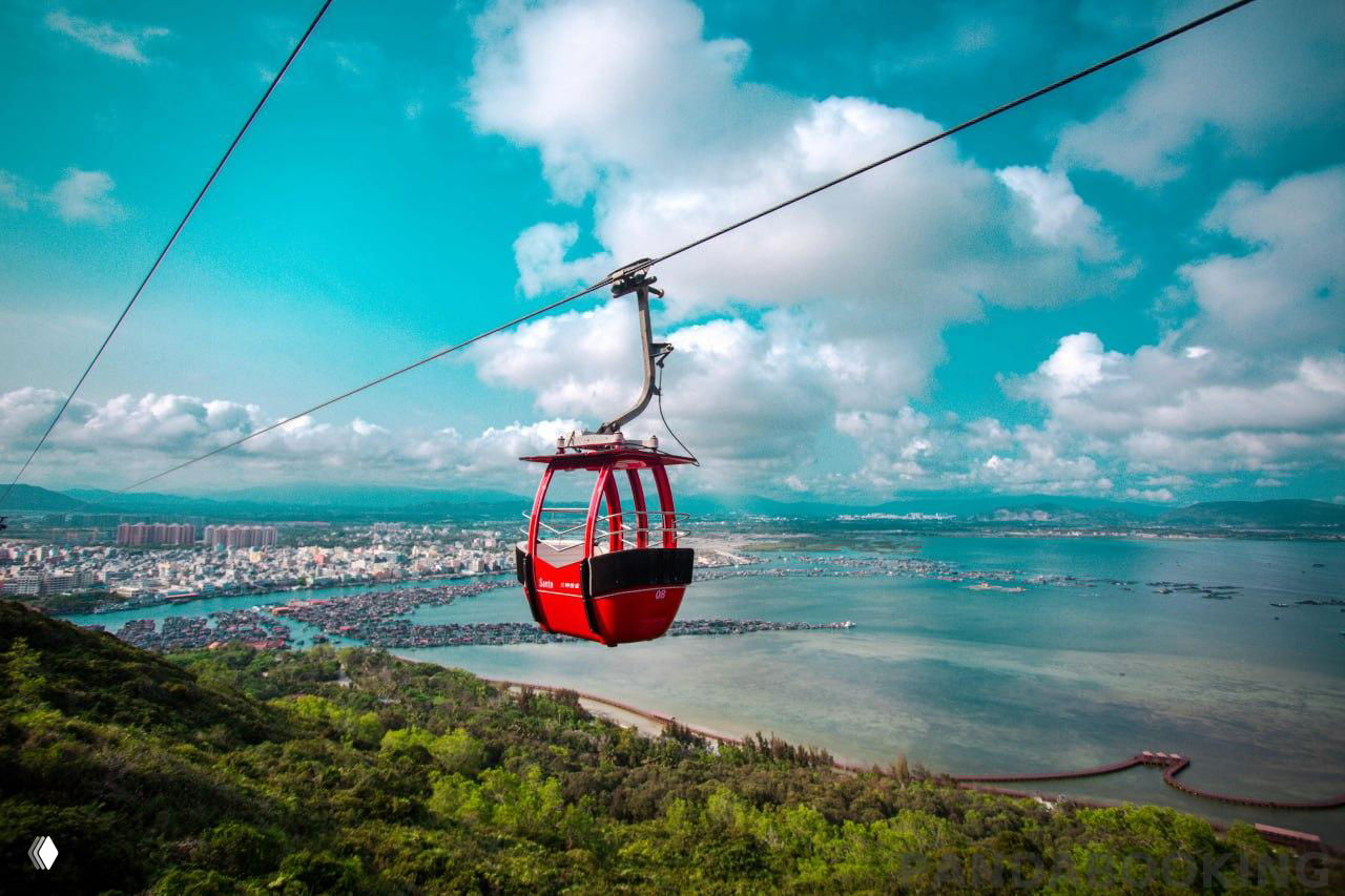 Красная кабинка фуникулёра над прибрежным холмом и морской гладью, вид на побережье и город Хайнань под облачным небом.