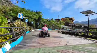 Туристическая деревянная дорожка в тропическом парке с маленьким электрокаром и пассажиром в розовой куртке, на фоне зелёных джунглей и строений.