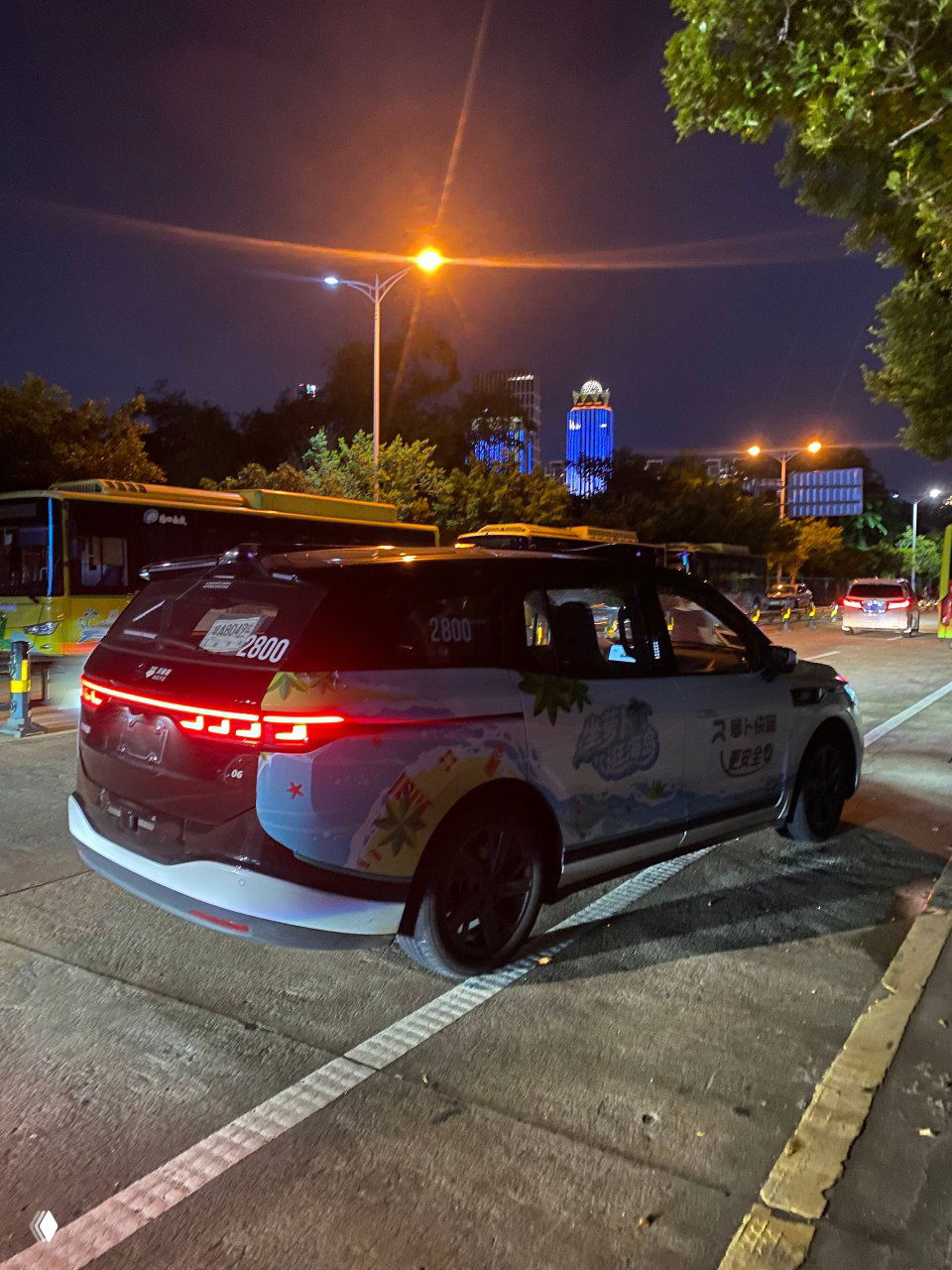 Ночное фото внешнего вида беспилотного такси Baidu Apollo в Хайкоу на городской стоянке с подсветкой и высотными зданиями на заднем плане, демонстрирующее дизайн кузова.