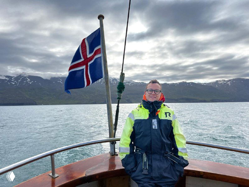 Пассажир на носу лодки в спасательном костюме на фоне фьорда и подчеркнуто развевающегося флага Исландии в надвигающейся облачности.