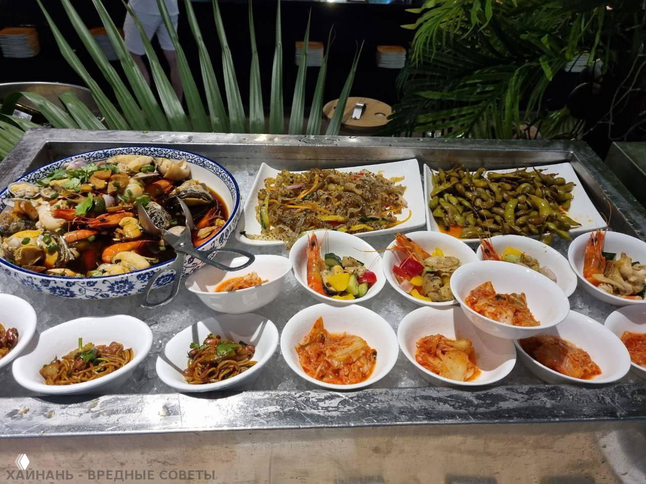 Небольшие белые миски с закусками, кимчи и морепродуктами, а также большое блюдо с рыбой и креветками на охлаждённом подносе шведского стола, общий вид ассортимента.