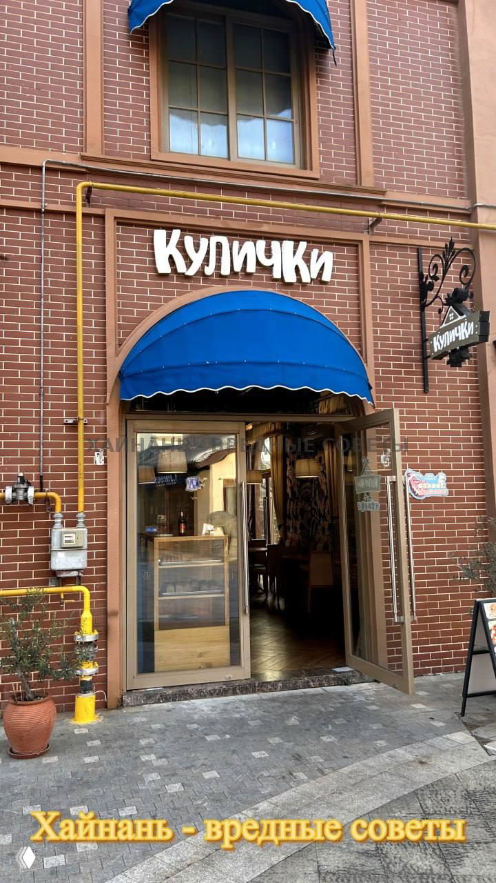 Фасад кафе «Куличики» на Цветочном острове: кирпичная стена, вывеска с названием, синяя маркиза и открытая дверь, видимо вход с тротуара.