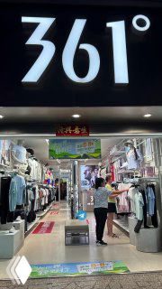 Вход в магазин 361 в Санье: крупная светящаяся вывеска «361°», видна витрина с одеждой и человек у входа на торговой улице.