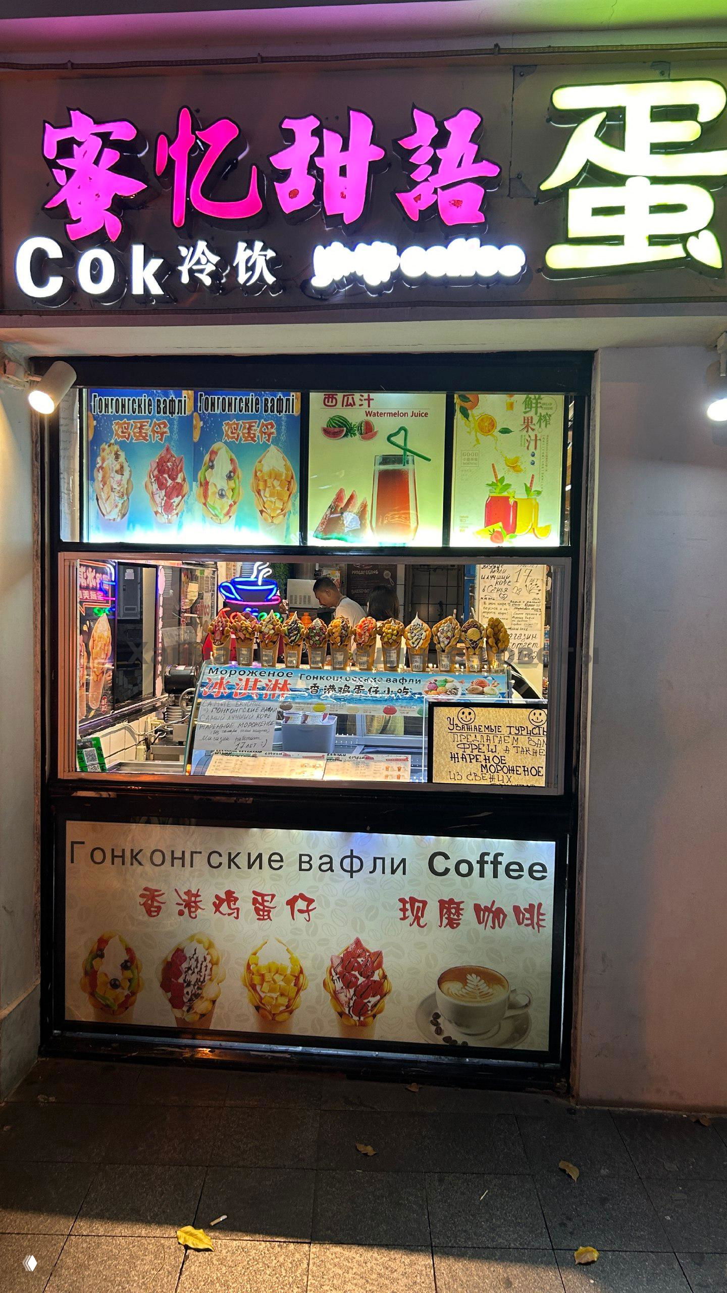Ночной фасад небольшого кафе с неоновой вывеской, витрина с ассортиментом гонконгских вафель и меню на стекле, видна русская и китайская вывеска.