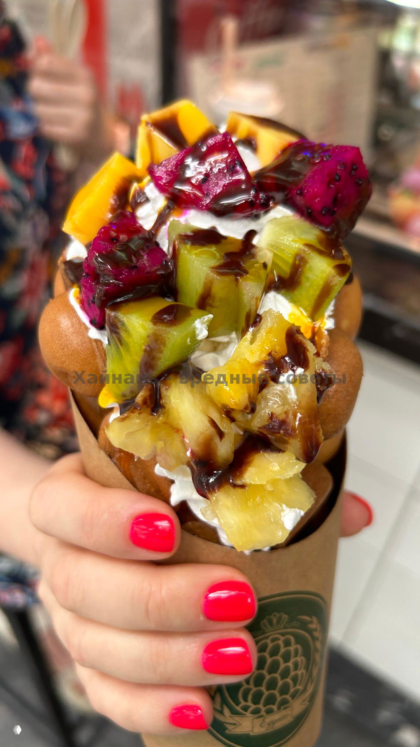 Крупный план гонконгской вафли в бумажной упаковке с кубиками драгонфрута, киви и ананаса, сверху тянется шоколадный соус, видна рука с розовым маникюром.