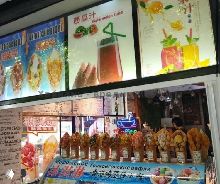 Широкий кадр маленькой уличной точки продаж с витриной, на переднем плане множество готовых вафель в стаканчиках, внутри видны продавцы и неоновая вывеска.