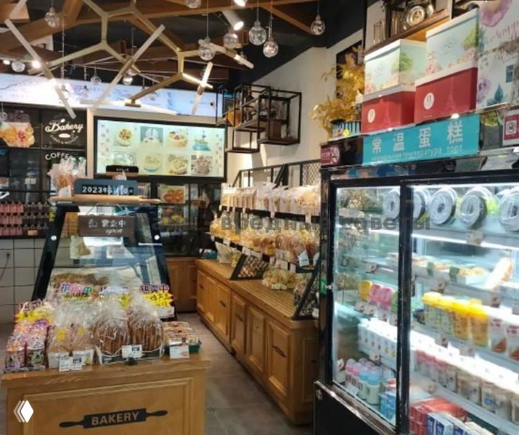 Интерьер мини-пекарни: деревянные прилавки с упакованной выпечкой, полки с товарами и большой холодильник с напитками и соками на фоне.