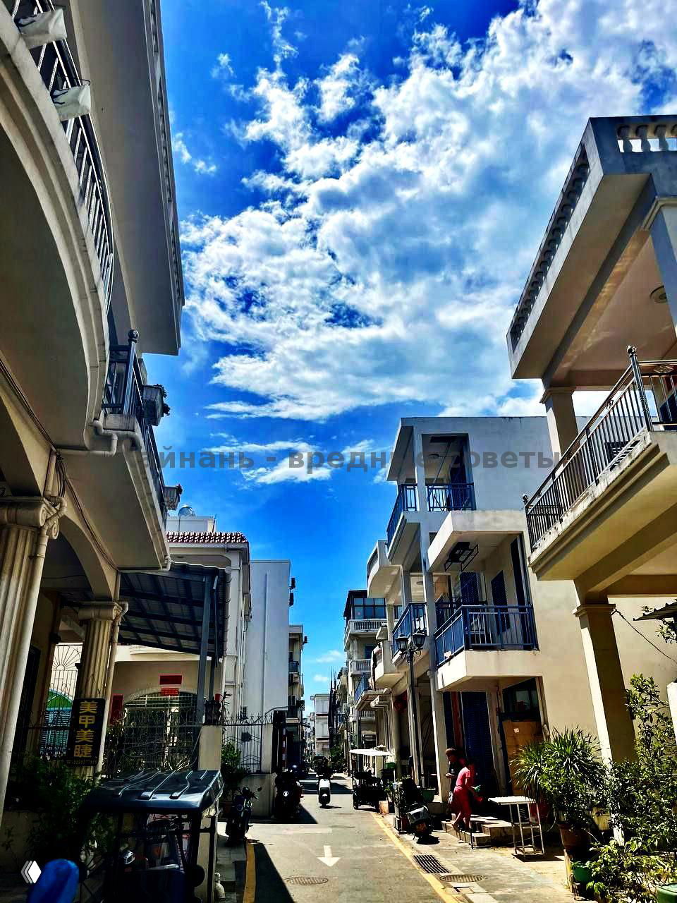 Узкая улица между белыми зданиями с балконами и парковочными скутерами под голубым небом — типичная городская застройка курортного квартала Танья.