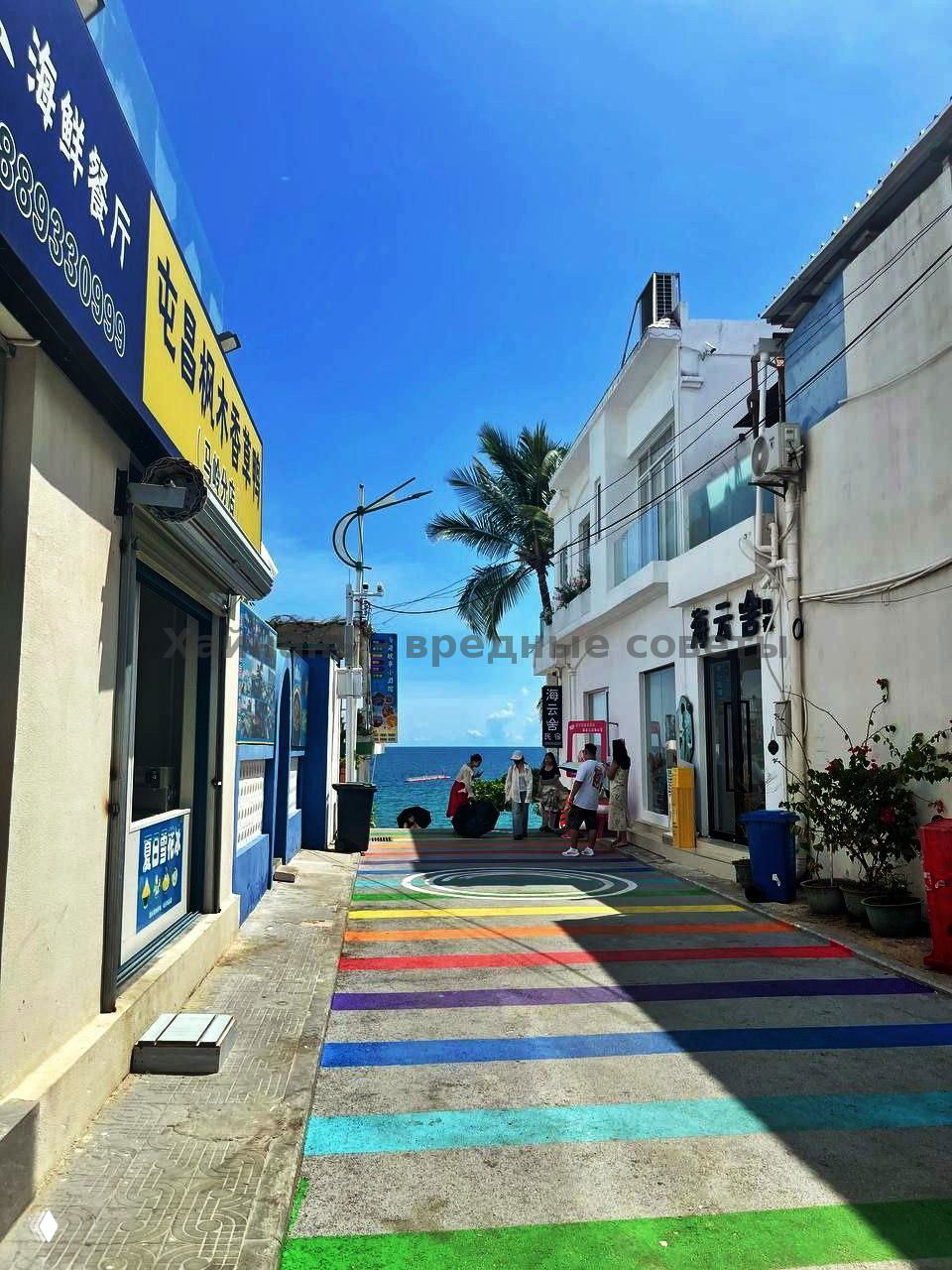 Цветная пешеходная дорожка, ведущая к морю: на конце видна кромка воды и пальмы, туристы подходят к берегу в солнечную погоду.