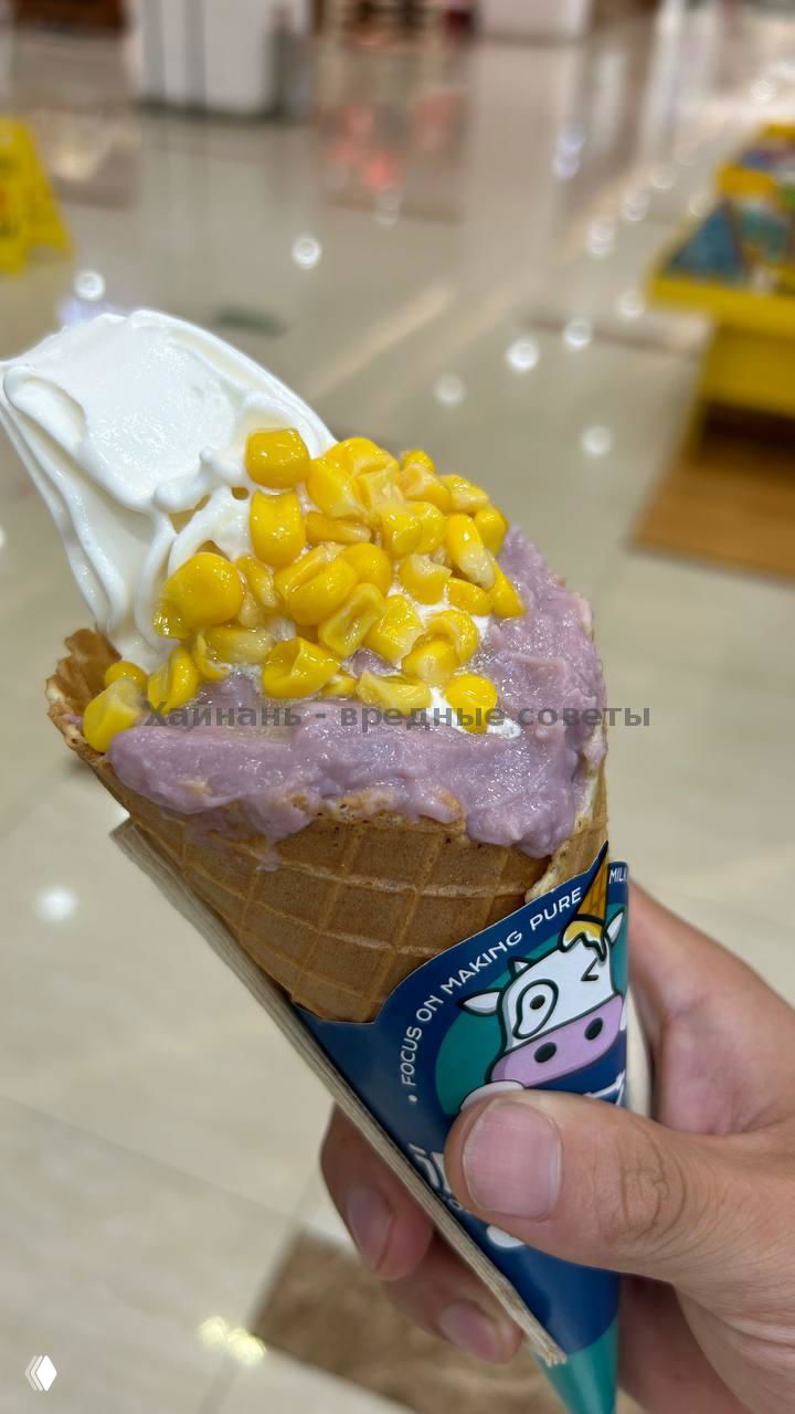 Крупный план вафельного рожка с белым мягким мороженым и крупными зернами кукурузы сверху; рожок держат в руке, видна рука держателя.