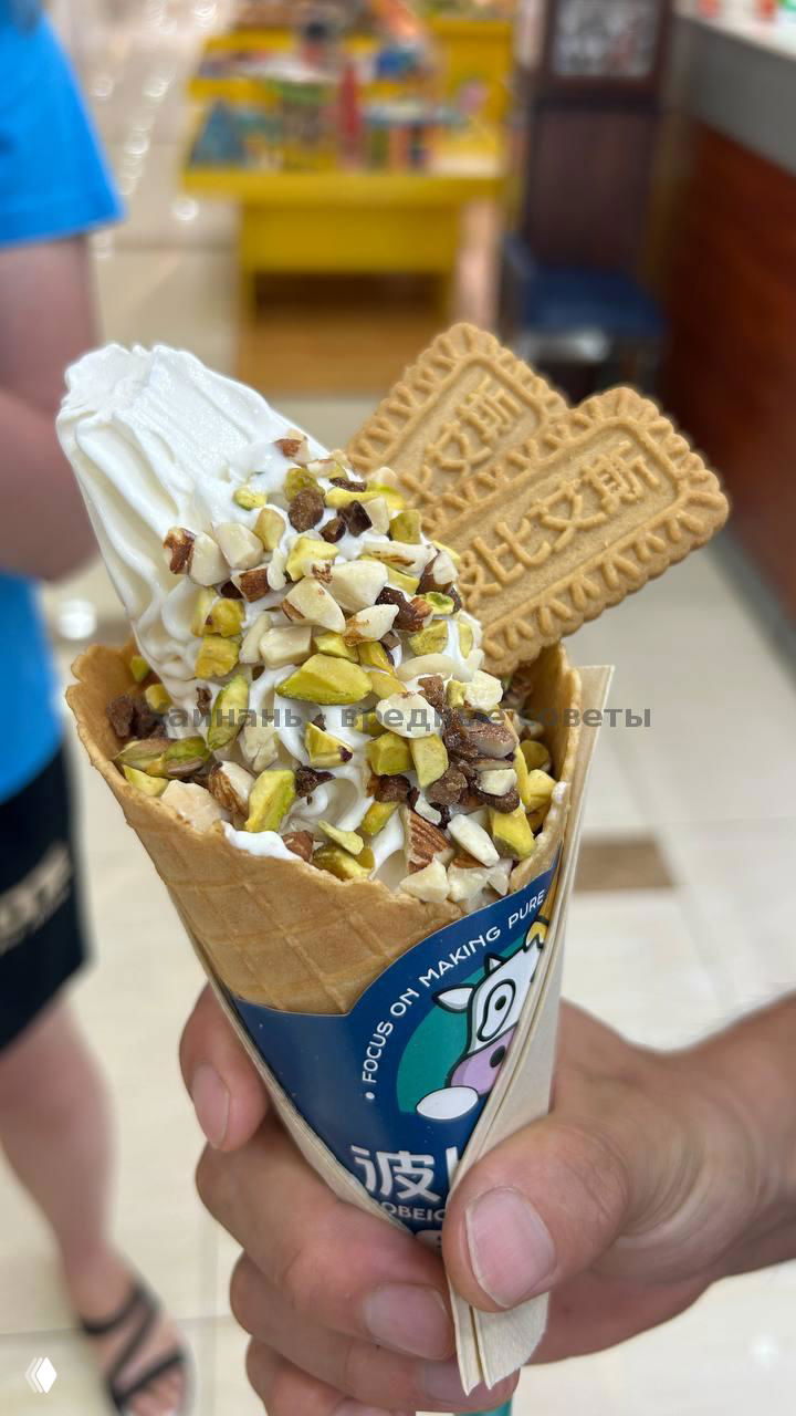 Вафельный рожок с мягким мороженым, рассыпанные дроблёные орехи и два декоративных печенья‑вкладыша, рожок держат в руке у прилавка.