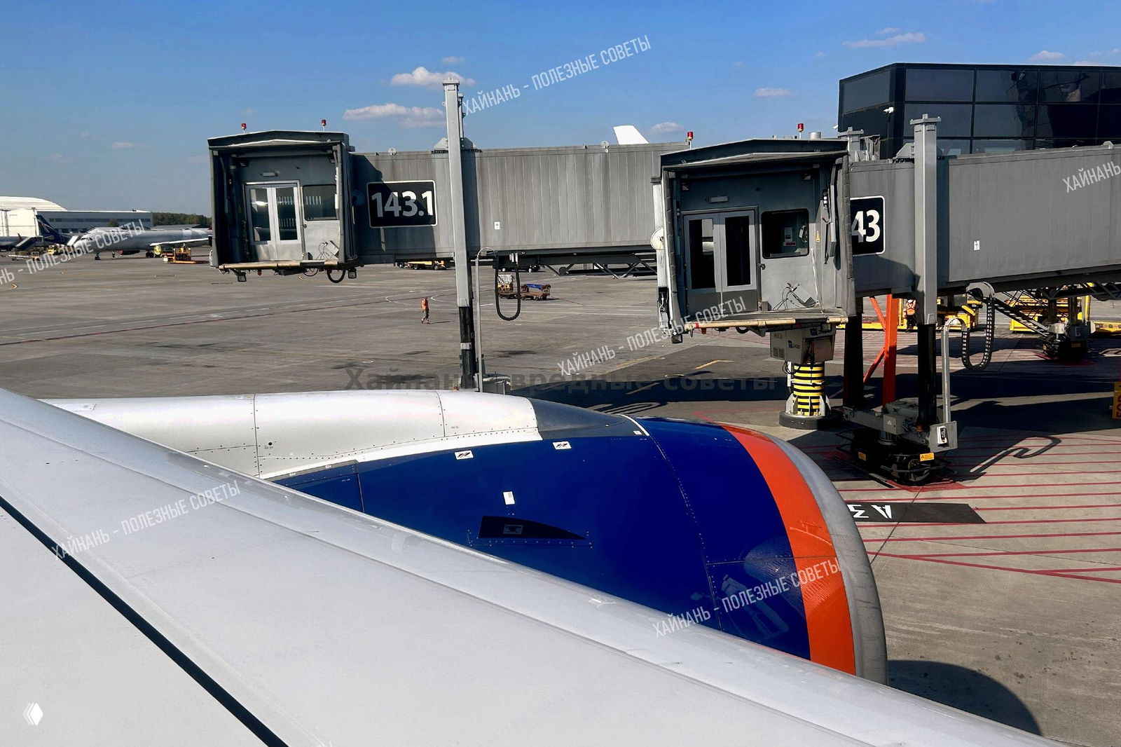 Вид из иллюминатора на двигатель самолёта и телетрап у гейта; видна часть крыла, рампа и терминал на фоне голубого неба.