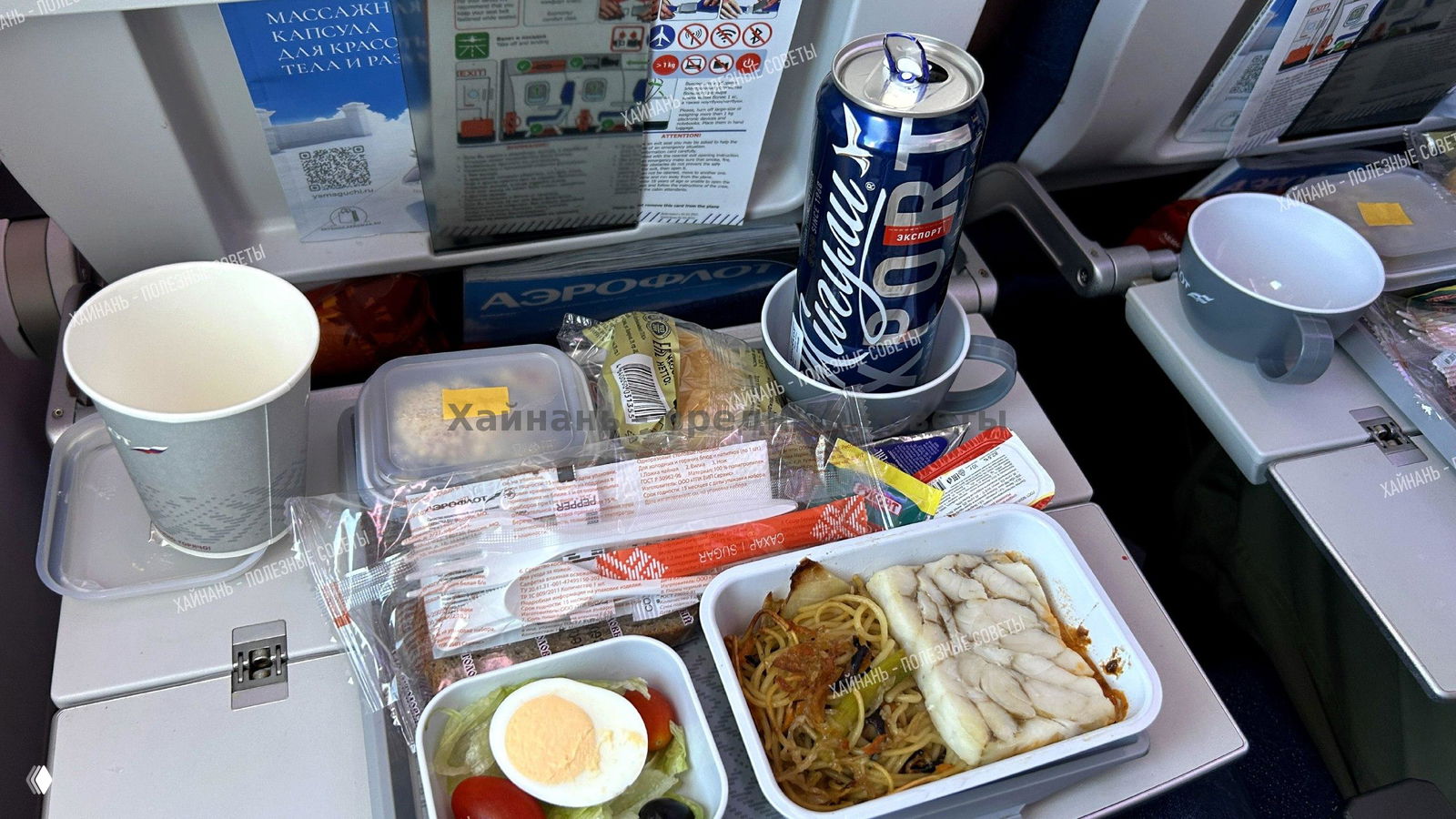 Поднос с бортовым питанием: салат, контейнер с основным блюдом, холодные закуски и банка пива на откидном столике.
