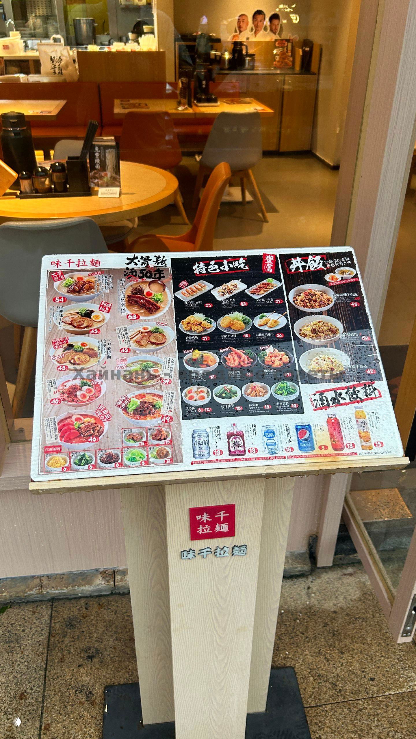 Стойка с меню у входа в AJISEN RAMEN: карточки с фотографиями блюд, разделы рамена, закусок и напитков с указанием цен в CNY.