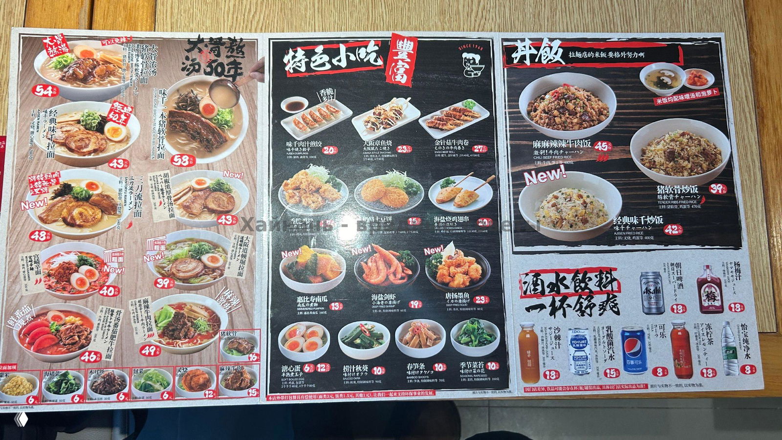 Развёрнутое меню на столе с крупными изображениями разных видов рамена, гарниров и закусок, снято сверху в ресторане.