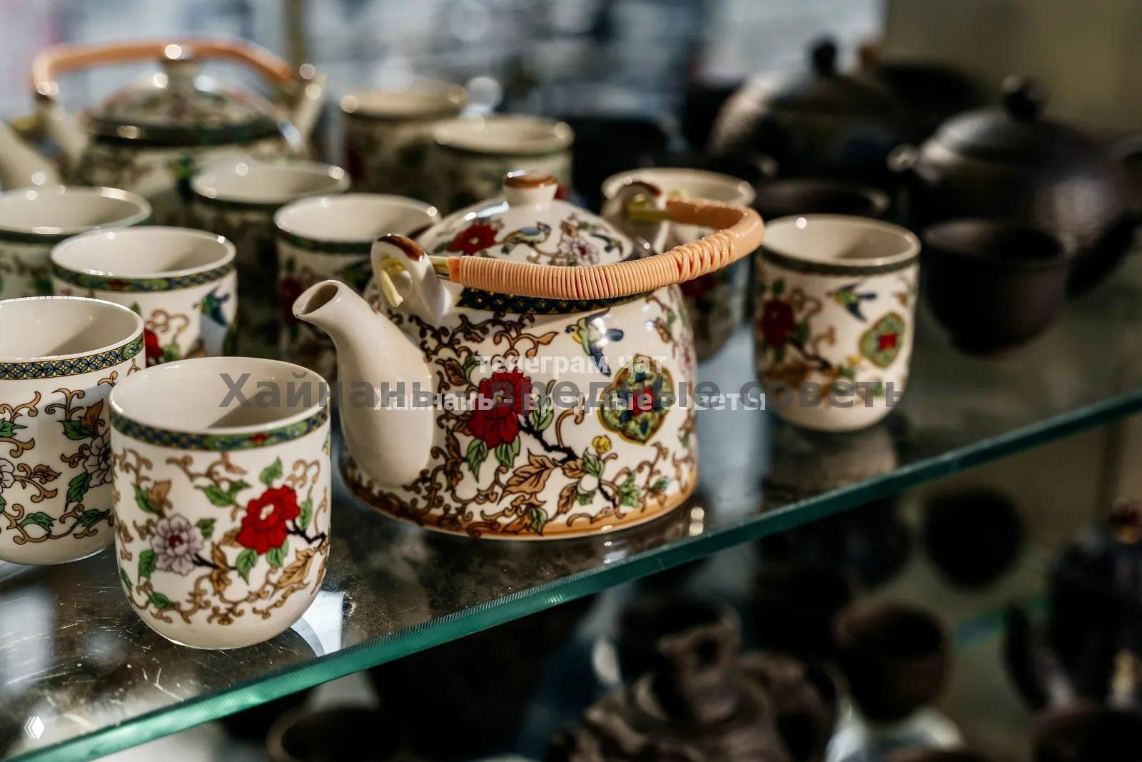 Крупный план чайной посуды и фарфорового чайника с узорами, расставленные на стеклянной полке среди других аксессуаров для чая.