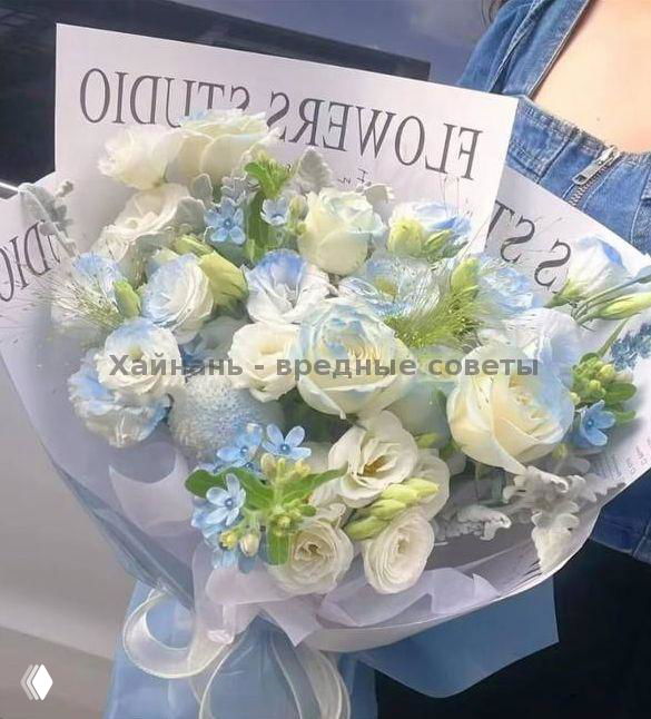 Пышный букет бело-голубых и кремовых цветов с зелёными акцентами в фирменной обёртке с логотипом FLOWERS STUDIO, держат на руках на фоне одежды.